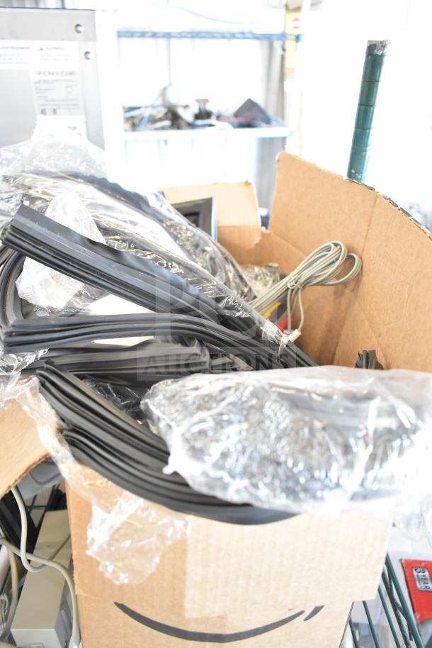 Lot of assorted cables and gaskets in a cardboard box, mostly black and gray, some in plastic wrapping, used condition.