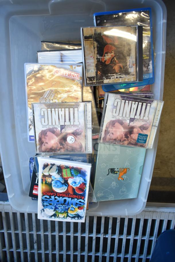 Lot of various items in a plastic bin, including DVDs like "Titanic," "Smurfs: A Christmas Carol," and assorted media.