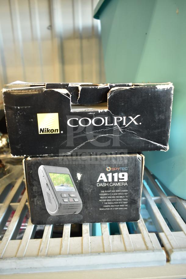 Nikon CoolPix digital camera box and SpyTec A119 dash camera box showing visible wear, on metal shelf.