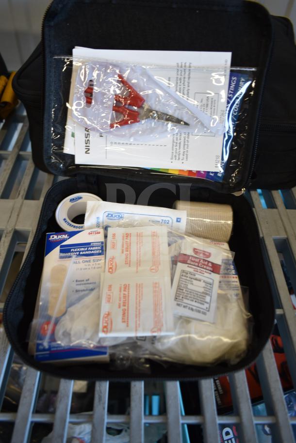 Nine assorted automotive kits including Hyundai, Nissan, Genesis, Kia first aid kits, and a Volkswagen roadside assistance kit, shown in a black zippered bag containing medical supplies such as bandages and cream.