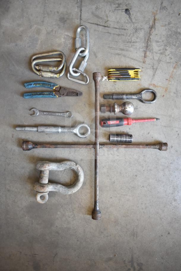 Assorted tools and hardware including carabiners, pliers, wrench, hitch ball, and screwdriver laid on concrete. Some rust visible.