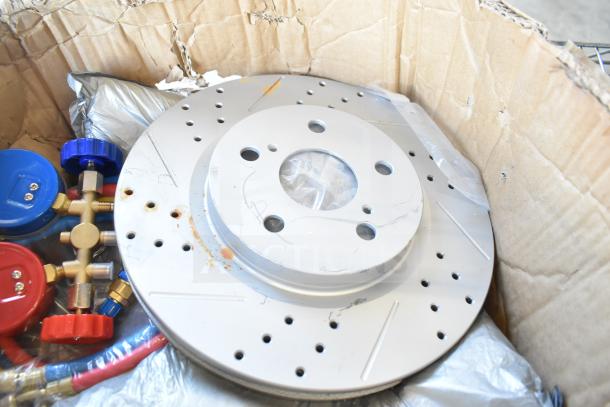 Lot of various items including gauges, hoses, and metal pieces in a cardboard box. Disc brake rotor visible on top. Mixed condition.
