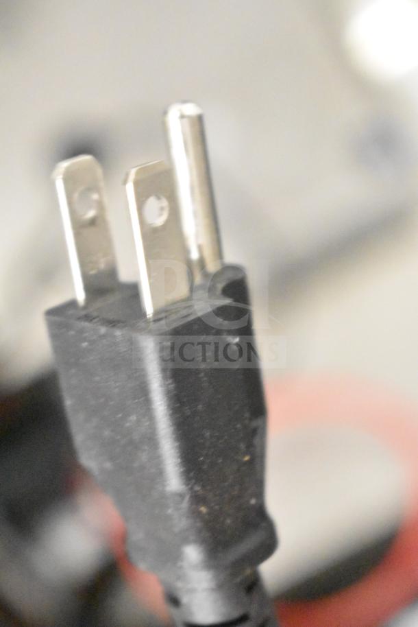 Close-up of a power plug from a mixed lot including a Kitchen Armor monitor, Netgear router, and PlayStation controllers.