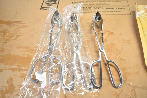 Three metal ice cream scoops with trigger handle in plastic wrap, on a cardboard surface.