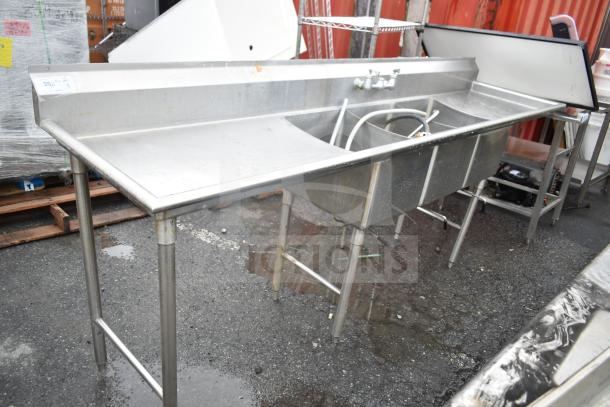 Commercial 3 bay sink with dual drain boards, stainless steel, used condition, visible faucet and plumbing attachments.