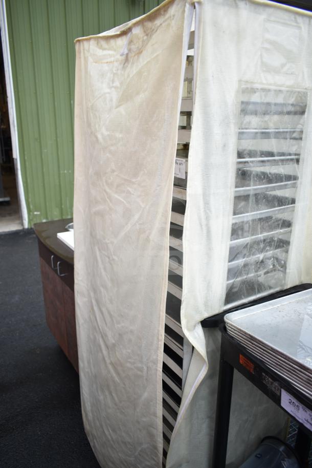 Pan transport rack with baking pans, covered with a protective drape. Visible wear on the cover. Multiple shelving units.