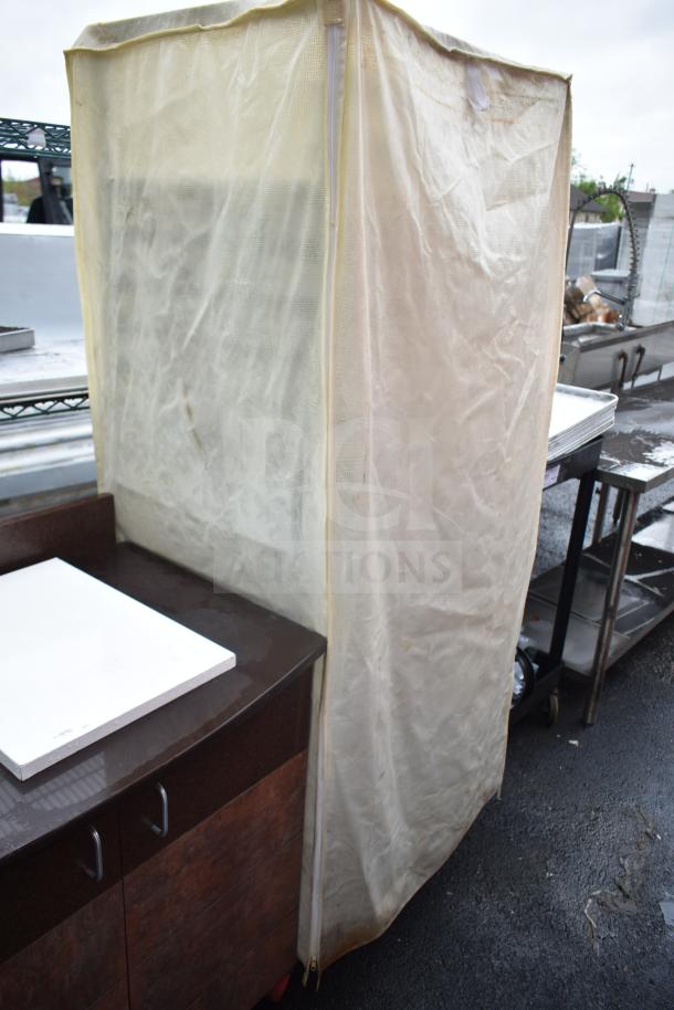 Pan transport rack with baking pans and protective cover. Cover is beige and zipped; condition appears used.