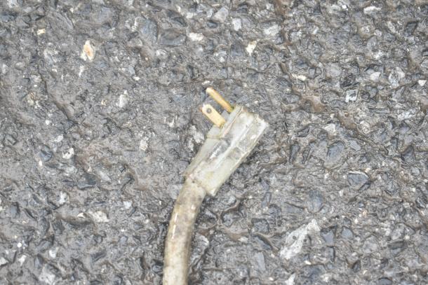 A frayed electrical power plug with exposed prongs lying on a wet asphalt surface.