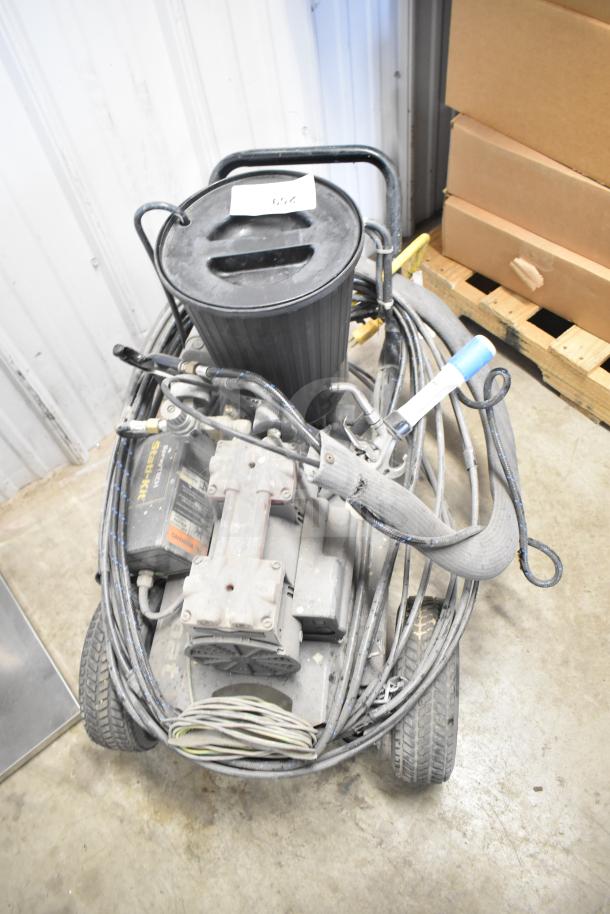 CapSpray compressor/pump with attached hoses and handle, showing wear, on wheels. Dusty surface, label partially visible.