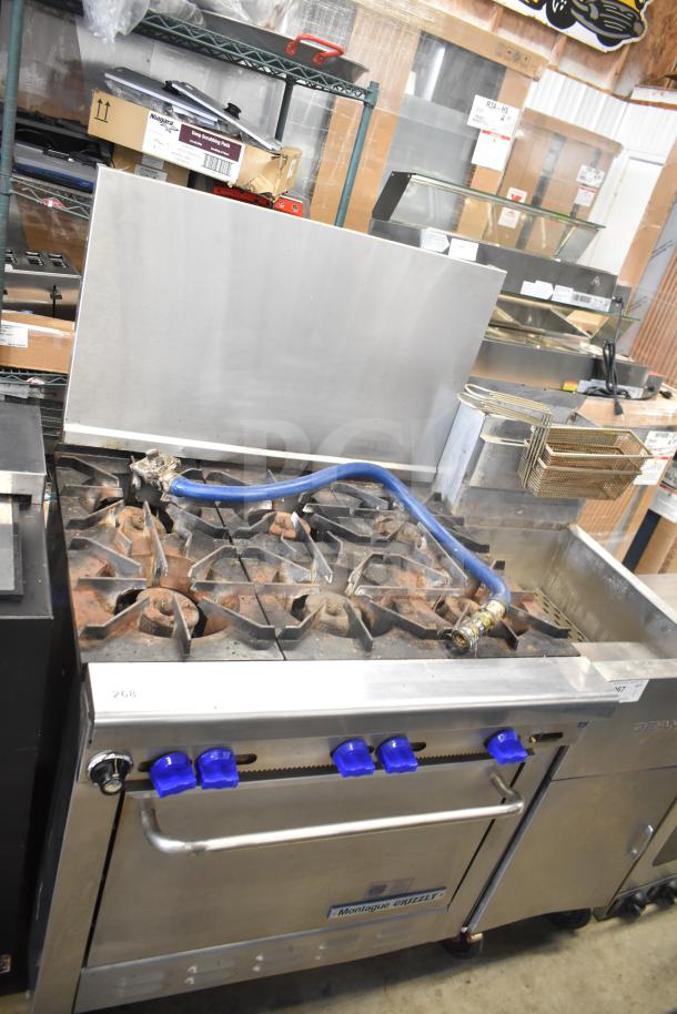 Montague Grizzly commercial 6-burner gas range with oven and backsplash, used condition, blue control knobs visible.