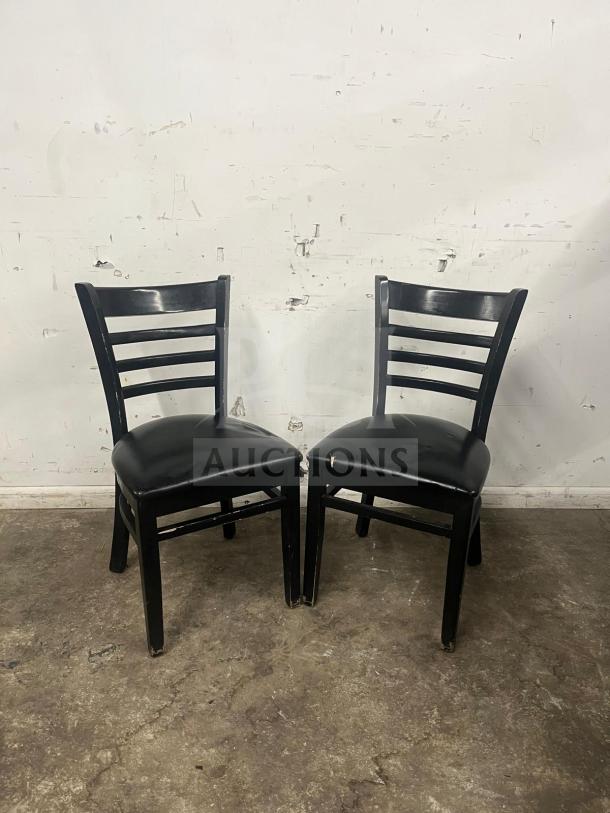 Two cafe dining chairs with black frames, padded vinyl seats, and ladder backs. Visible wear on legs.
