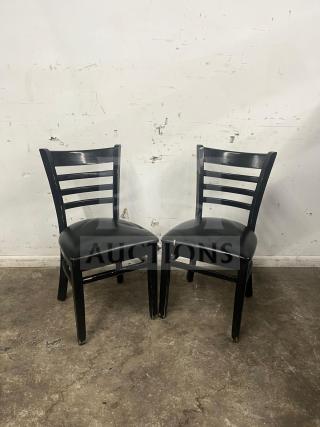 Two cafe dining chairs with black frames, padded vinyl seats, and ladder backs. Visible wear on legs.