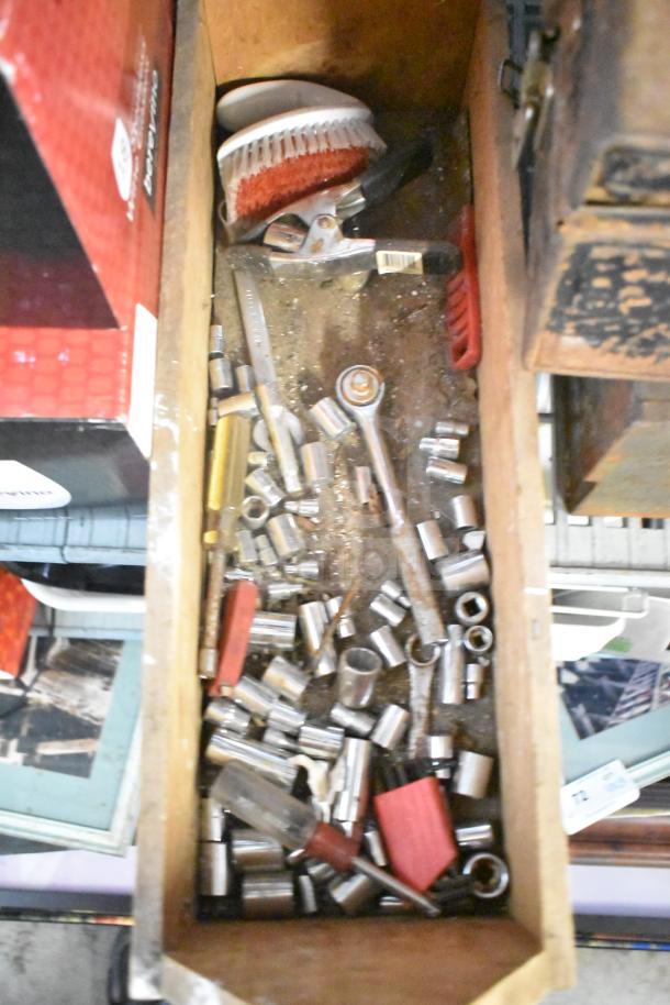 Assorted tools including a ratchet, sockets, clamps, and a scrub brush in a wooden box. Items show signs of use.
