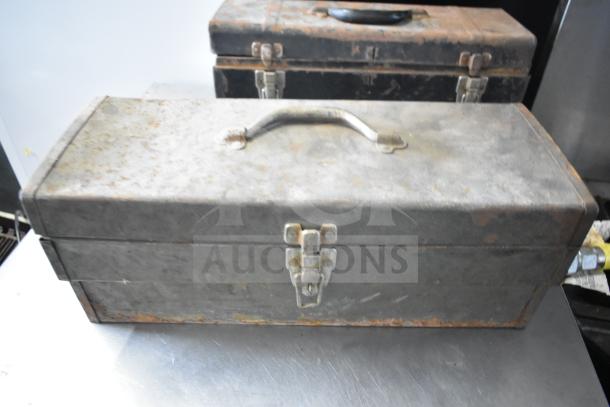 Rustic metal toolbox with handle and latches; aged, weathered condition. Includes clamps, jumper cables, tools.
