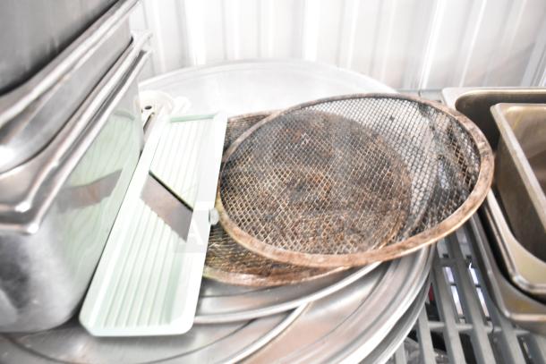 Lot of kitchen items including stainless steel bins, green mandoline slicer, mesh strainers, and round baking sheets.