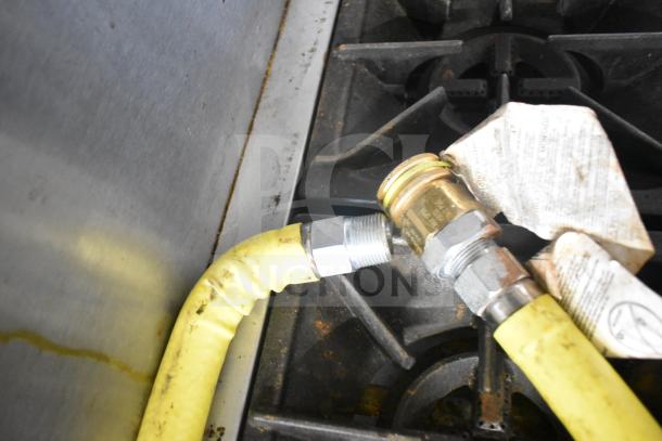 A close-up view of a yellow gas line connection attached to a commercial range burner. Tags and fittings visible.
