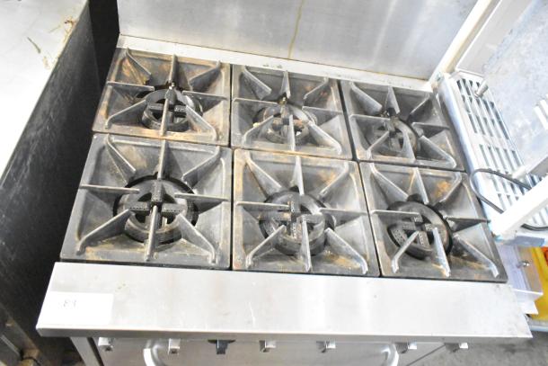 Commercial gas-powered 6-burner range with oven, over shelf, and backsplash. Slight wear, model number visible on bottom left.