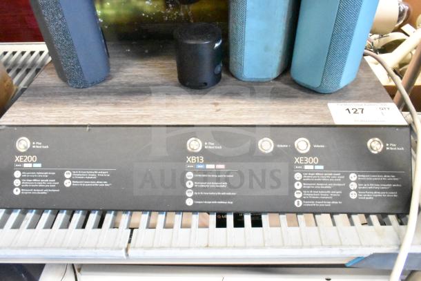 Speaker store display featuring three portable Bluetooth speakers: XE200, XB13, and XE300. Includes play and volume controls, with product descriptions highlighting features like waterproof design and battery life. Auction tag labeled "127."