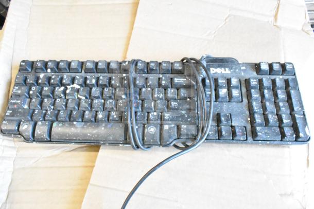 Dell computer keyboard with visible wear and dust, resting on cardboard. Features standard QWERTY layout and USB cable.