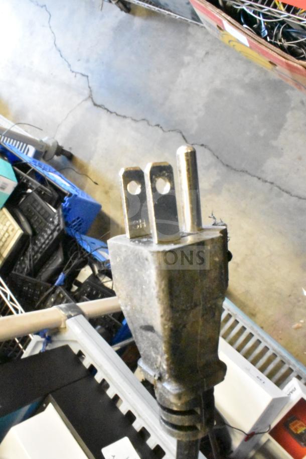 Power plug close-up from a Soundcore store display with speakers, showing wear. Plug marked "SH."