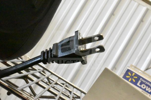 Black speaker with attached power cord, featuring a standard two-prong plug. Mounted on a metal shelf.