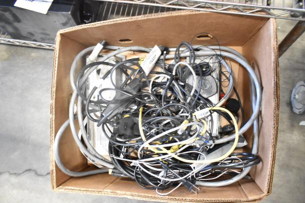 Box of mixed cables and electronics, including Sensormatic ZBSTPSP-L. Varied condition, potentially valuable components.