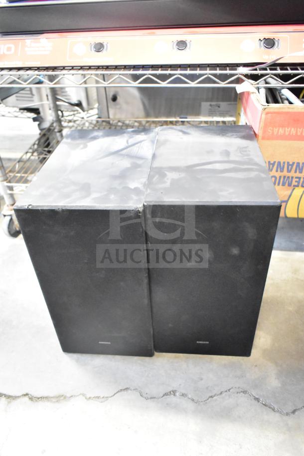 Two Samsung PS-WB65D wireless subwoofers, black, in good condition, placed on a metal shelf.