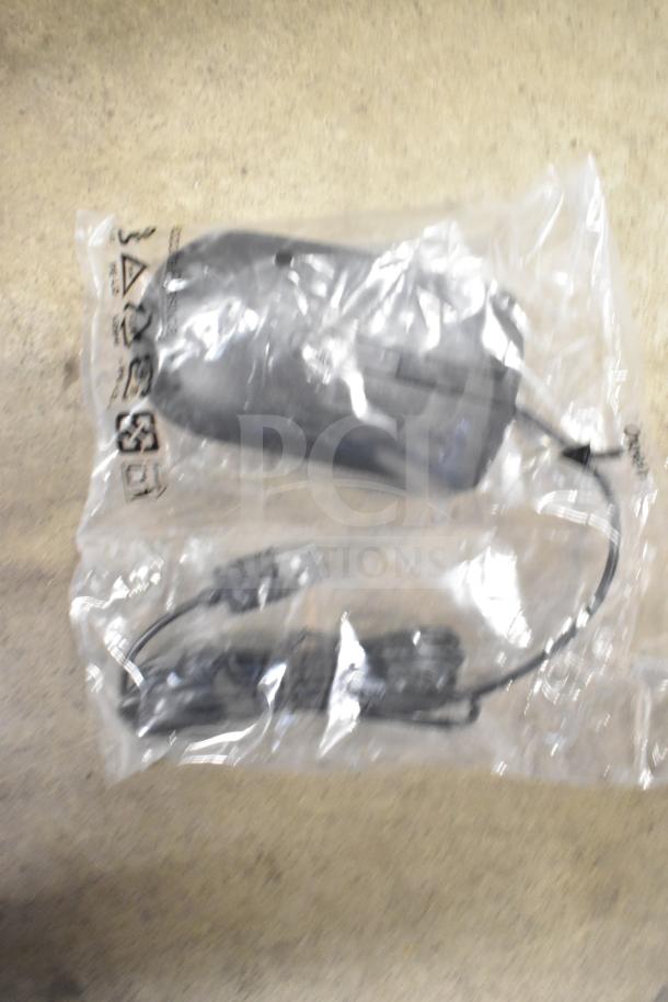 Sealed package containing a computer mouse and cables, with visible recycling symbol and clear plastic wrap. 