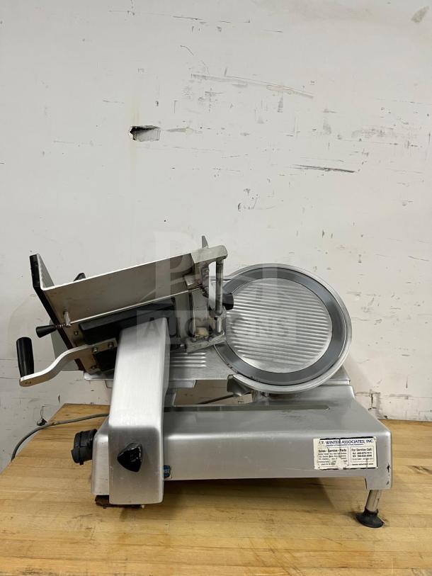 Steel manual food slicer on a wooden surface, featuring a circular blade and handle. Label indicates manufacturer details. Good condition.