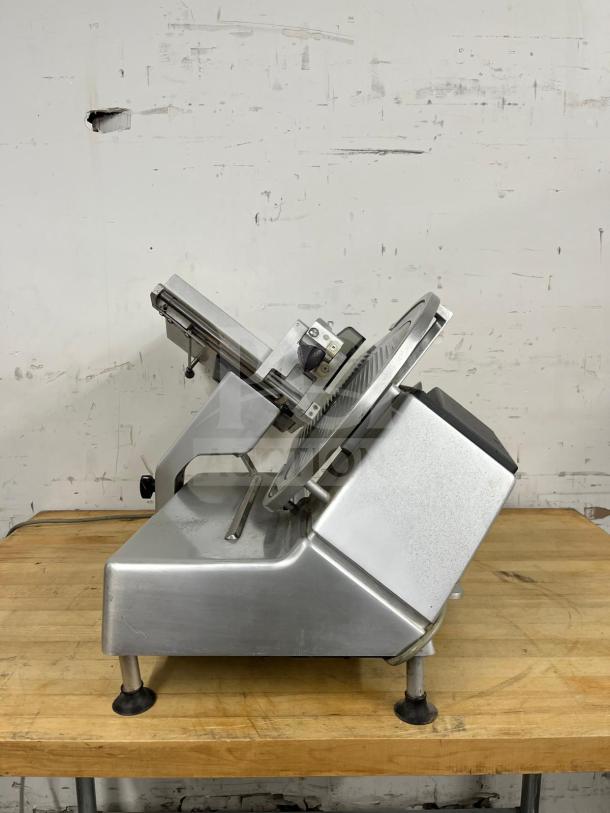 Steel manual food slicer on wooden table, showing blade and guard. In good condition with minor wear. Ideal for precise slicing.