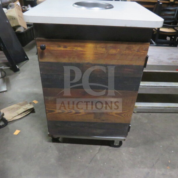 One-door trash receptacle on casters, wood finish, metal top with waste hole, dimensions 27x27x40 inches, used condition.