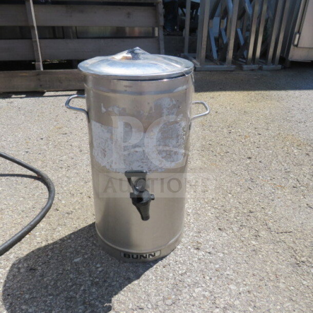 Bunn stainless steel satellite with lid, visible wear, side handles, and spout; brand label on the bottom front.