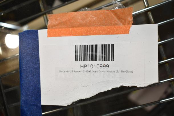 Barcoded label for Garland/US Range oven door window, model HP1010999, on metal surface, taped with blue and orange tape.