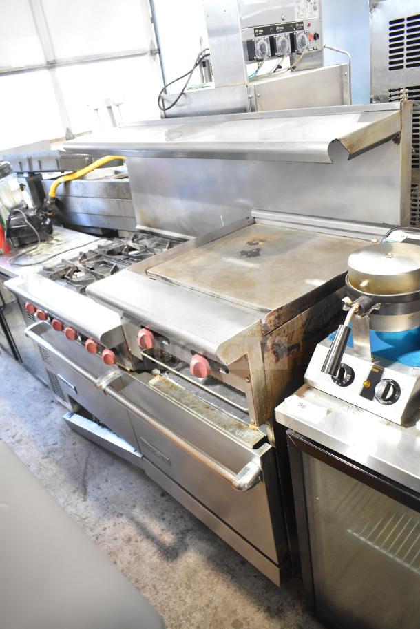 American Range commercial 6 burner range with flat top, 2 ovens, over shelf, and backsplash. Visible wear, sturdy condition.