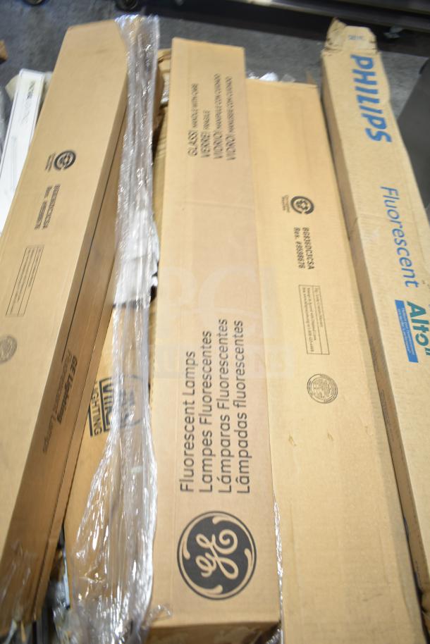 Boxes of fluorescent tube lights, brands include GE and Philips, with visible scratch and dent condition, unopened packaging.