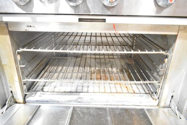 Commercial gas-powered 6-burner range with oven, overhead shelf, and backsplash. Visible wear, sturdy construction.