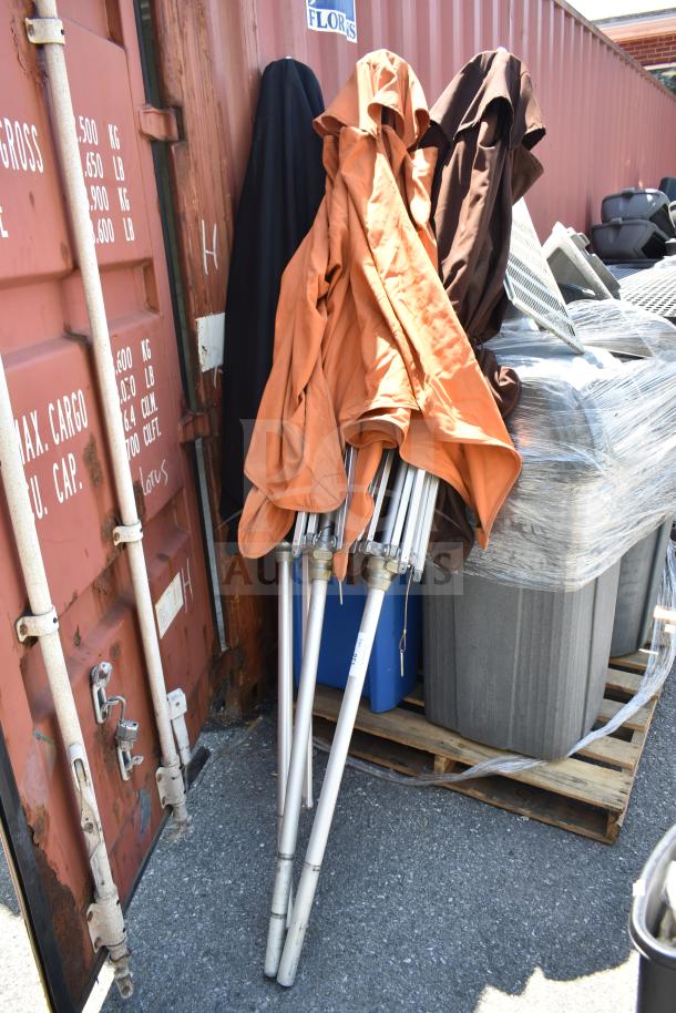 Three patio umbrellas, varied colors, folded with metal frames. Used condition, resting against a shipping container.