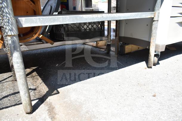 Commercial metal table with under shelf, sturdy legs, and visible caster wheels; outdoor use condition.