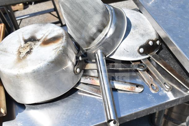 Stainless steel commercial table with under shelf, includes pot, strainer, and kitchen utensils, showing visible wear.