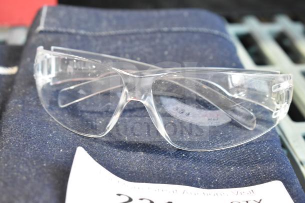 Brand new safety glasses with clear lenses on denim fabric, part of scratch and dent set including apron and gloves.
