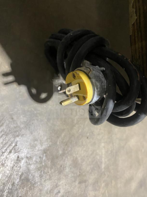 A coiled black power cord with a yellow pronged plug on a concrete surface.