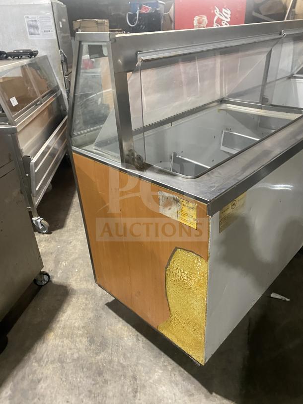 Commercial stainless steel ice cream dipping cabinet with slanted glass and rear lift-open access. Visible wear on the wood panel.