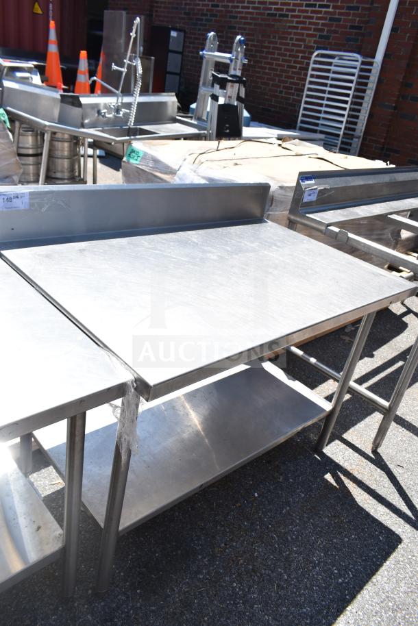 Stainless steel commercial table with backsplash and under shelf, used condition, visible tags indicating item details.