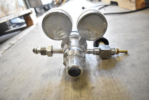 Heavy-duty metal gauge with two dials, showing connectors and fittings, in used condition on concrete floor.