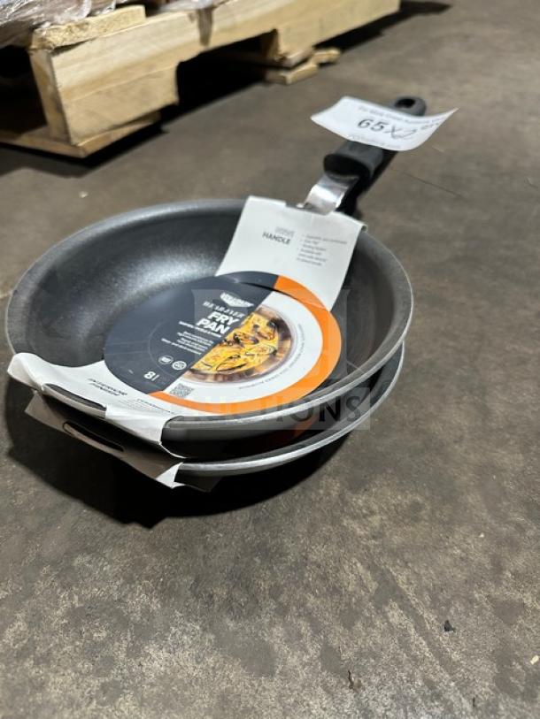 Stacked Vollrath frying pans with labels indicating size and handle details, on warehouse floor. New condition.