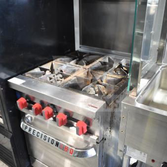 Precision commercial 4-burner gas range with oven, overshelf, backsplash, on casters. Visible wear, red control knobs.