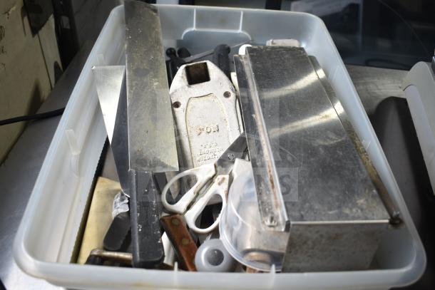 Plastic bin with assorted items including serving spoons, scissors, a metal box, and various tools, visibly worn.