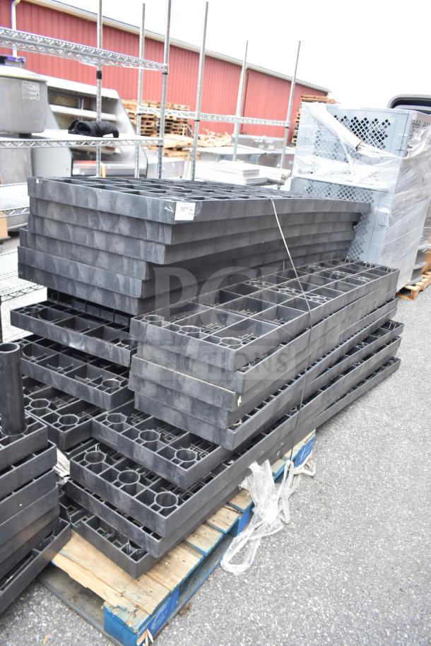 Alt text: Lot of 23 black poly shelf pieces stacked outdoors. Used condition, multiple square compartments per piece, on wooden pallets.