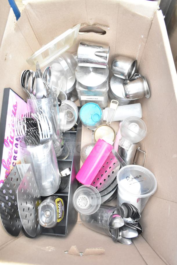 Cardboard box containing assorted kitchen items including cutlery, metal cups, plastic lids, and labeled containers. Mixed condition.
