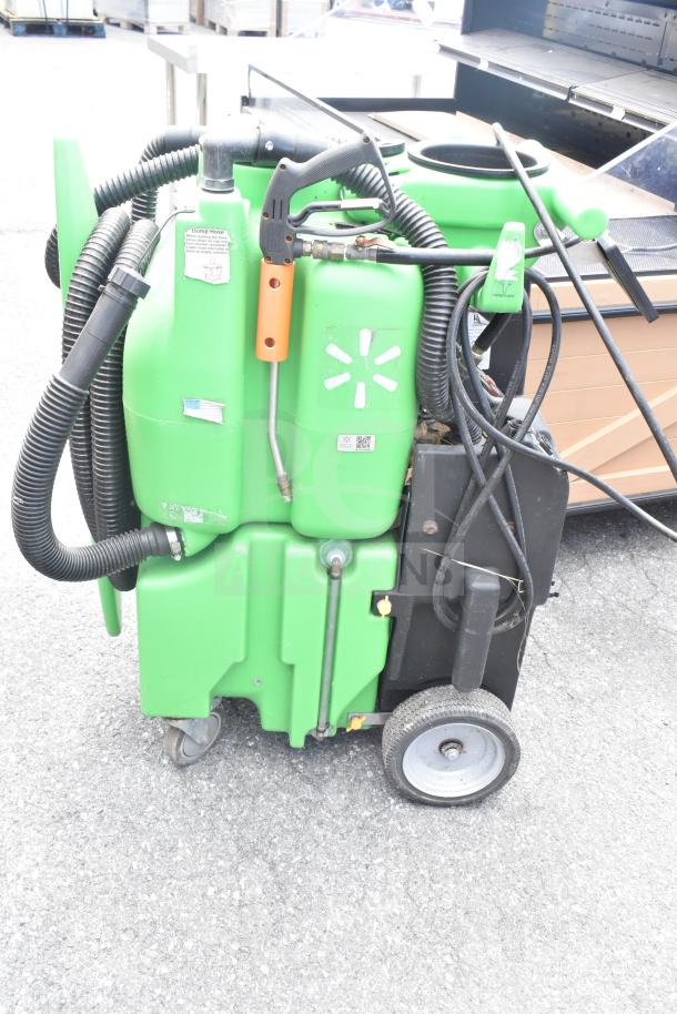 Green floor cleaning machine on wheels with hoses and control handle, labeled with symbols, in used condition.